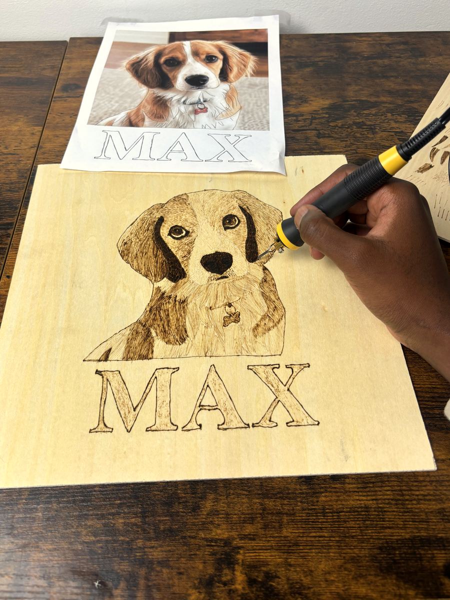A hand burning the final details of a fluffy dog portrait into a live-edge wood slab, with the name MAX burned below